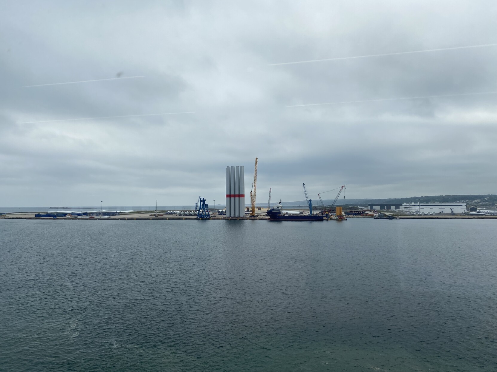 Grey harbour and sky with wind turbine blades and towers stacked on the quayside.