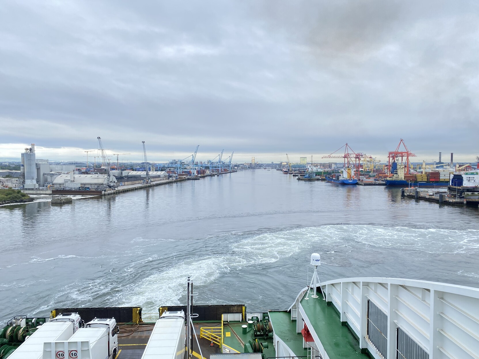 View from Irish Ferries Ulysses of the the river Liffey on departure from Dublin.  Some truck on the rear deck in the foreground. Grey river and engine churned waters in lower mid ground.  Port cranes and ships and Dublin skyline topped by our beloved leaden grey skies.  Forty shades of grey