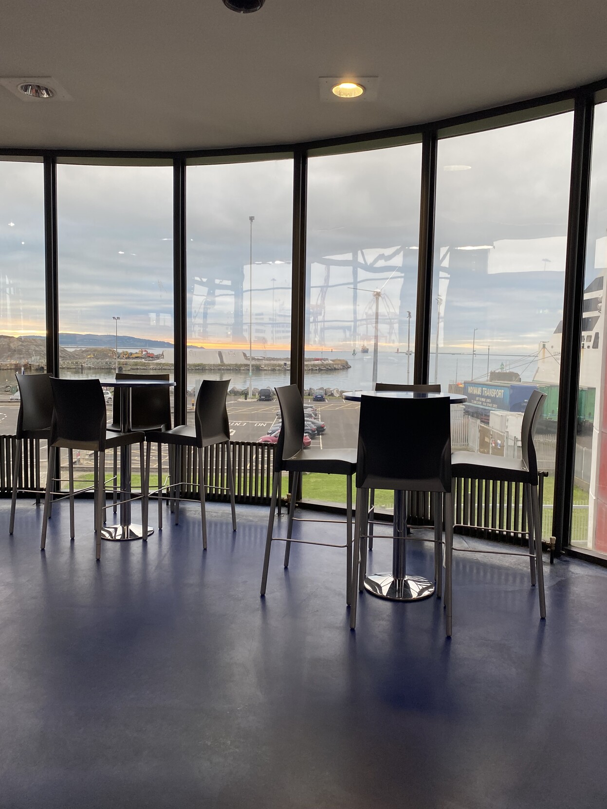 High tables and chairs in cafe beside floor to ceiling windows that open onto the port. 