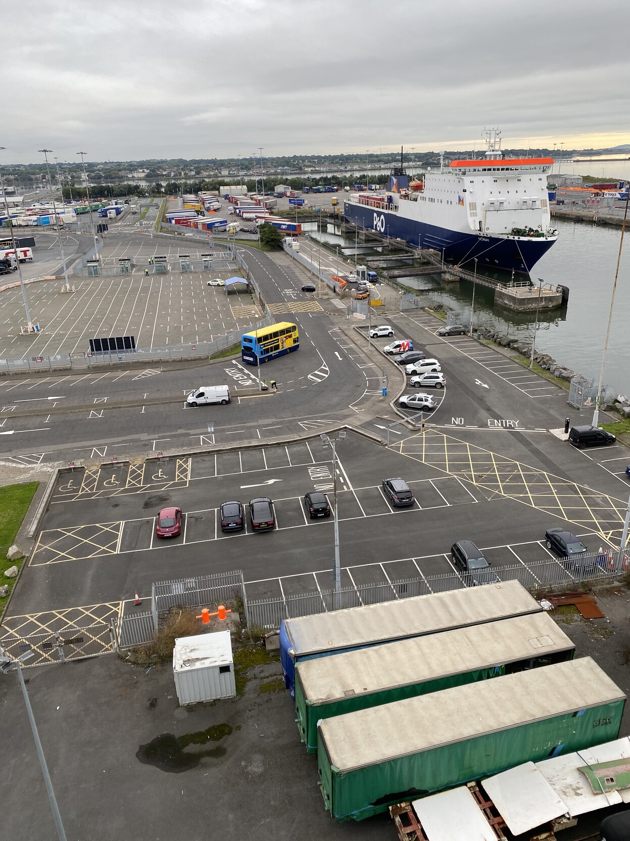 View of small car park from deck of ferry. Car park is a dog leg shape around the terminal approach road. There are a small number of parked cars and other traffic on roads and boarding lanes. The P&O ferry is also at berth and in view in the background