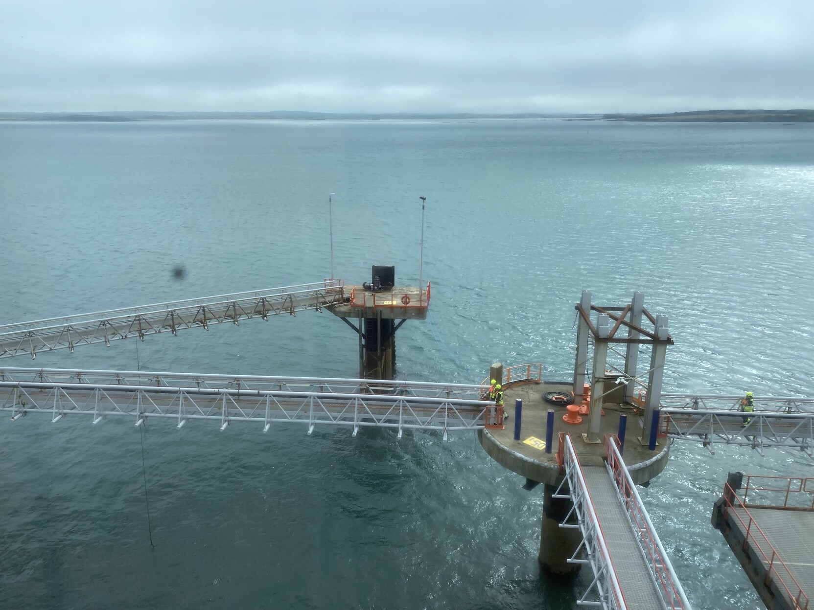 View from ferry window as we wait for the call for foot passengers. Pictured is a dock with narrow service walkways perched on columns sitting in the water. A little brightness on a generally green-grey sea