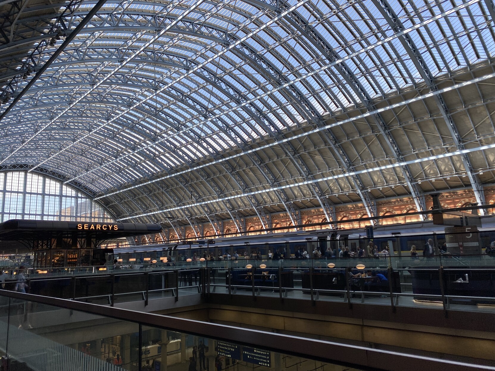 Bright sun drenched iron and glass roof of St Pancras station dominates the photo. A section of light grey roofing is terminated with bright orange and yellow brick walls forming a long series of closed arches. The lower third is darker and on close inspection a Eurostar train with passengers milling alongside is visible. Closer still a mostly empty champagne bar with the “Searcys” name prominent in white signage is perched above a glimpse of the lower level (in every way) of the station 