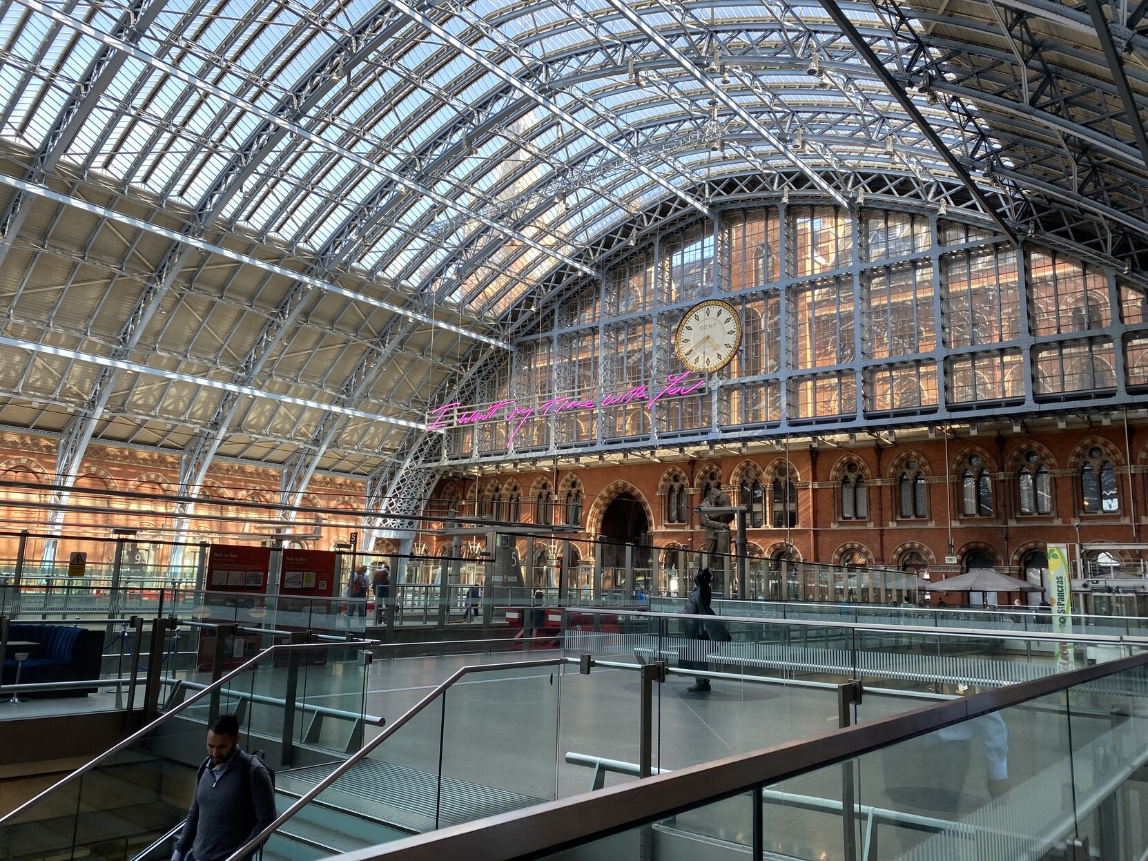 Looking back at the old entrance to St Pancras. The beautiful roof, described in the previous picture, ends in a glass wall that gives a glimpse of the orange and cream brick Victorian extravaganza of a station building. On the wall hangs a huge elegant gold and white clock face reads 4:40 (pm) and the make in black lettering is “Dent”. On the lower left in front of the glass a bright modern sign composed of individual neon pink cursive lettering reads “I want my time with you”. Below the glass element of the wall are two stories of high arched windows and to the left within these windows is a two story entrance arch. In the centre in front of the orange wall is a bronze? modern statue of a wartime couple in a farewell embrace. The majority of the foreground consists of the greys of modern glass, steel and flooring. Though just visible is a second statue of John Betjeman who led a defence of the station when plans were afoot to tear it all down in the name of progress.