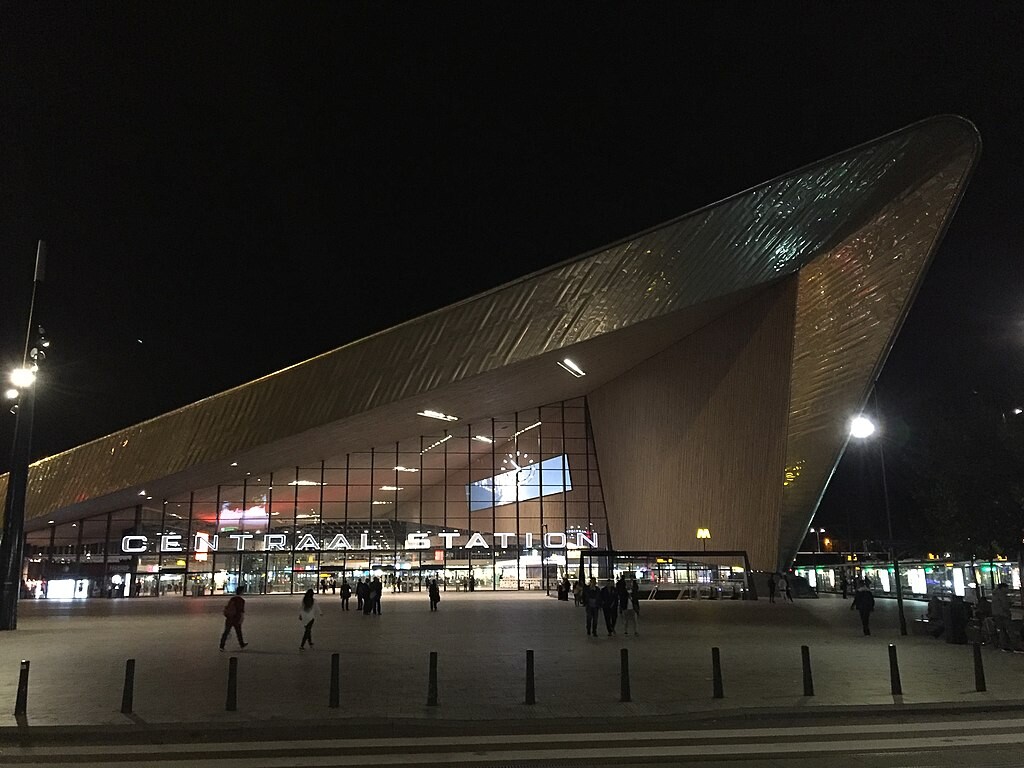 The spectacular front view of Rotterdam Centraal station at night. A wedge of grey roof projects outwards to meet a thick wedge of wall projecting outwards in tandem. The design screams movement. The glass wall of the station is recessed underneath. The original signage of the previous building is a beautiful statement in illuminated white in the bottom third of the glass wall. The station forecourt is sparsely populated with people.  Creative Commons Attribution 2.0 Generic License Author: https://www.flickr.com/people/21160385@N02