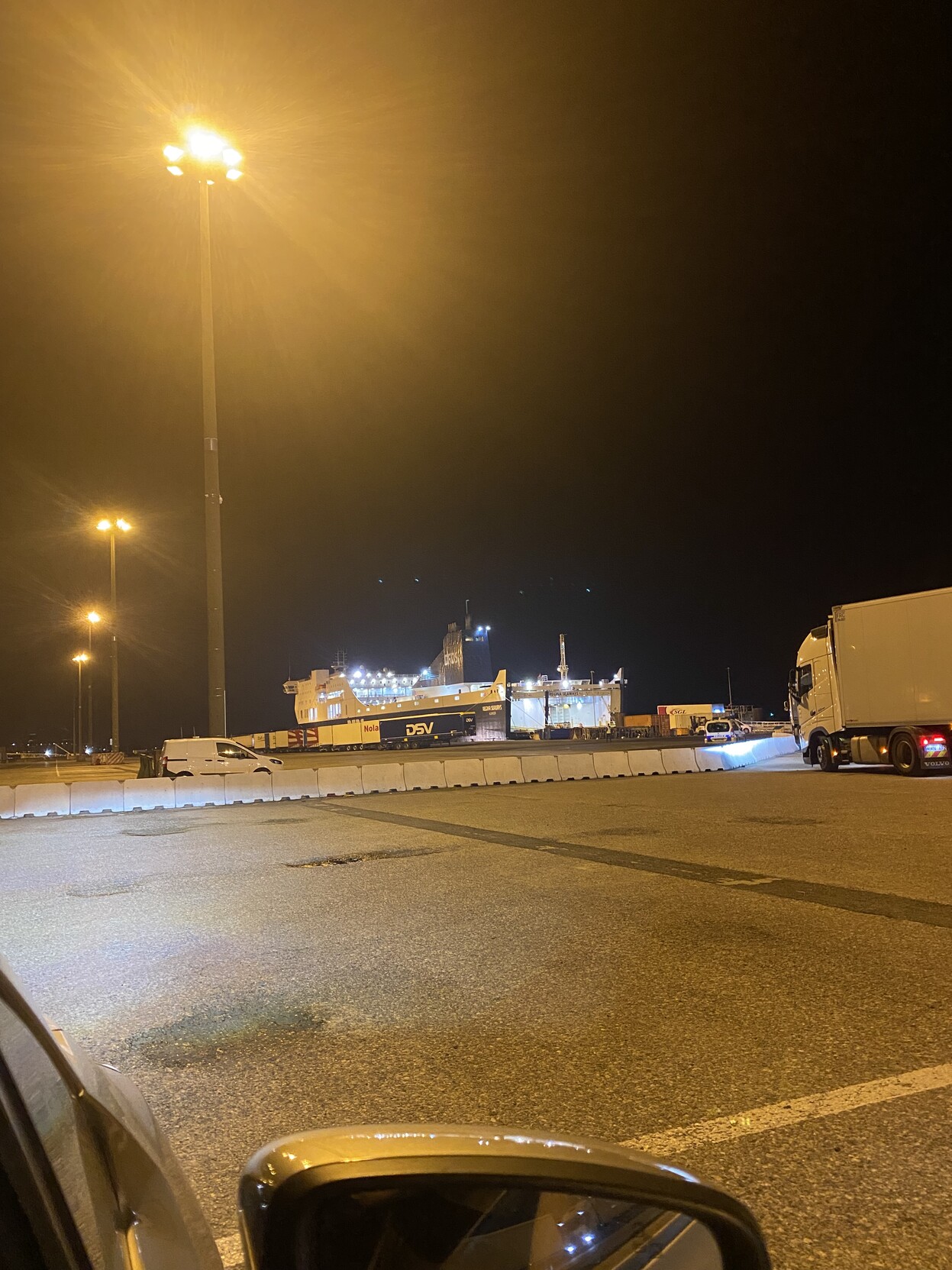 Small ferry in centre frame as viewed from a car with top of wing mirror in shot. Paved surface and concrete lane divider illuminated in yellow by lights on a tall light pole. Front of a articulated truck also in shot right of centre.