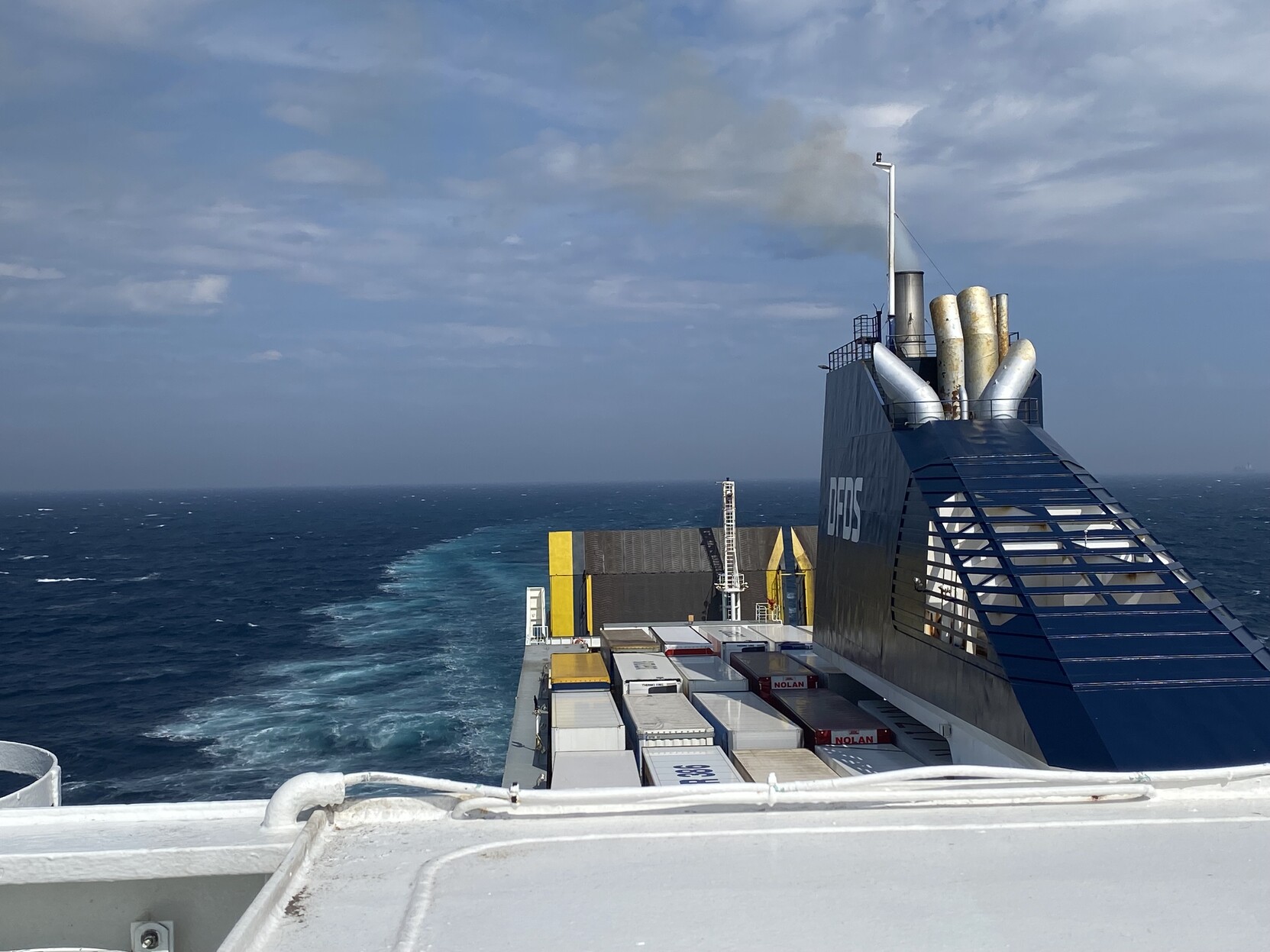 View from top deck of ferry looking back towards the stern. Mostly white truck trailers are packed on an open deck with the grey/brown rear ramp with bright yellow highlighted sections in a vertical closed position. The dark blue engine funnel is on the right of picture with DFDS in white lettering. The white wake of the ship in an inky blue sea is visible on the left and a bright partially cloudy sky rises from the distant horizon.