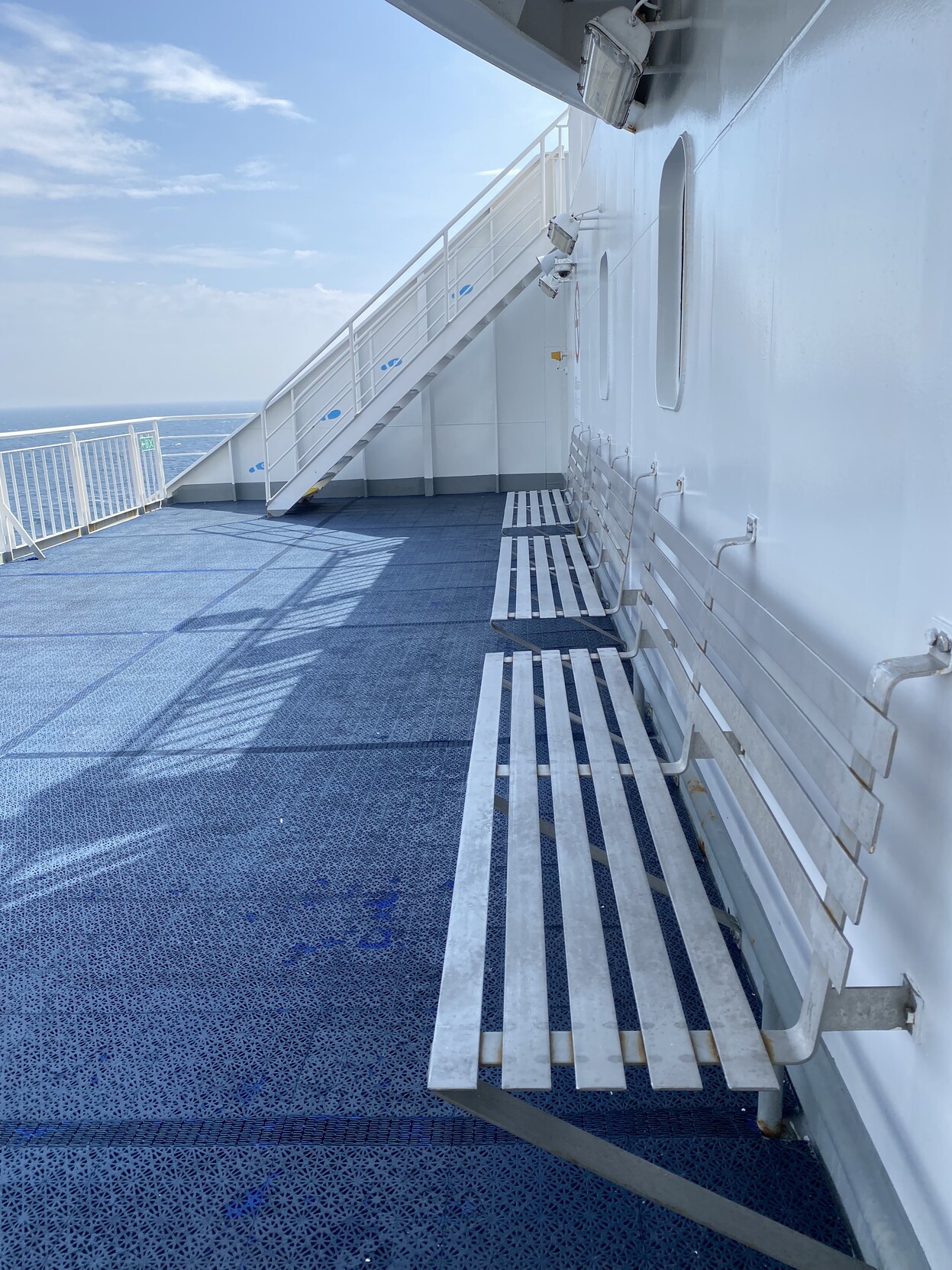 Open rear-facing small deck with a white wall, light grey metal benches and a bright blue rubber/plastic floor. The top right hand side contains a white metal railing and a bright partially cloudy sky. A white stairway to an upper deck runs from left to right at the rear centre of the field of view.