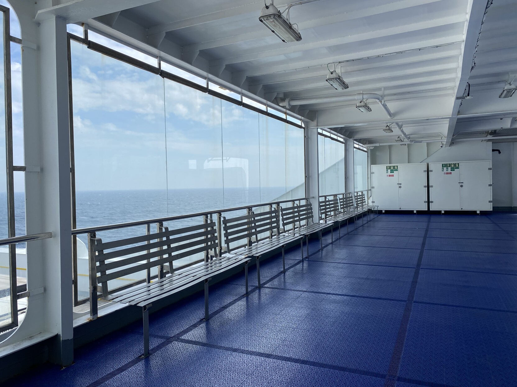 Rear facing deck mostly enclosed with a glazed wall that is faced with light grey metal benches. The floor is bright blue and the ceiling is a bright white