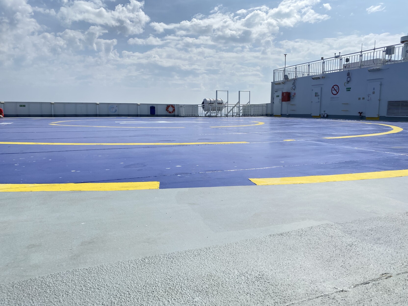 Open “sun deck” with helicopter landing spot marked in bright blue and yellow. The deck is painted light grey in the foreground outside the helicopter landing area. A white closed rail is in the mid distance and a white wall containing white doors to a further forward deck. Above the sky is bright and partially cloudy.