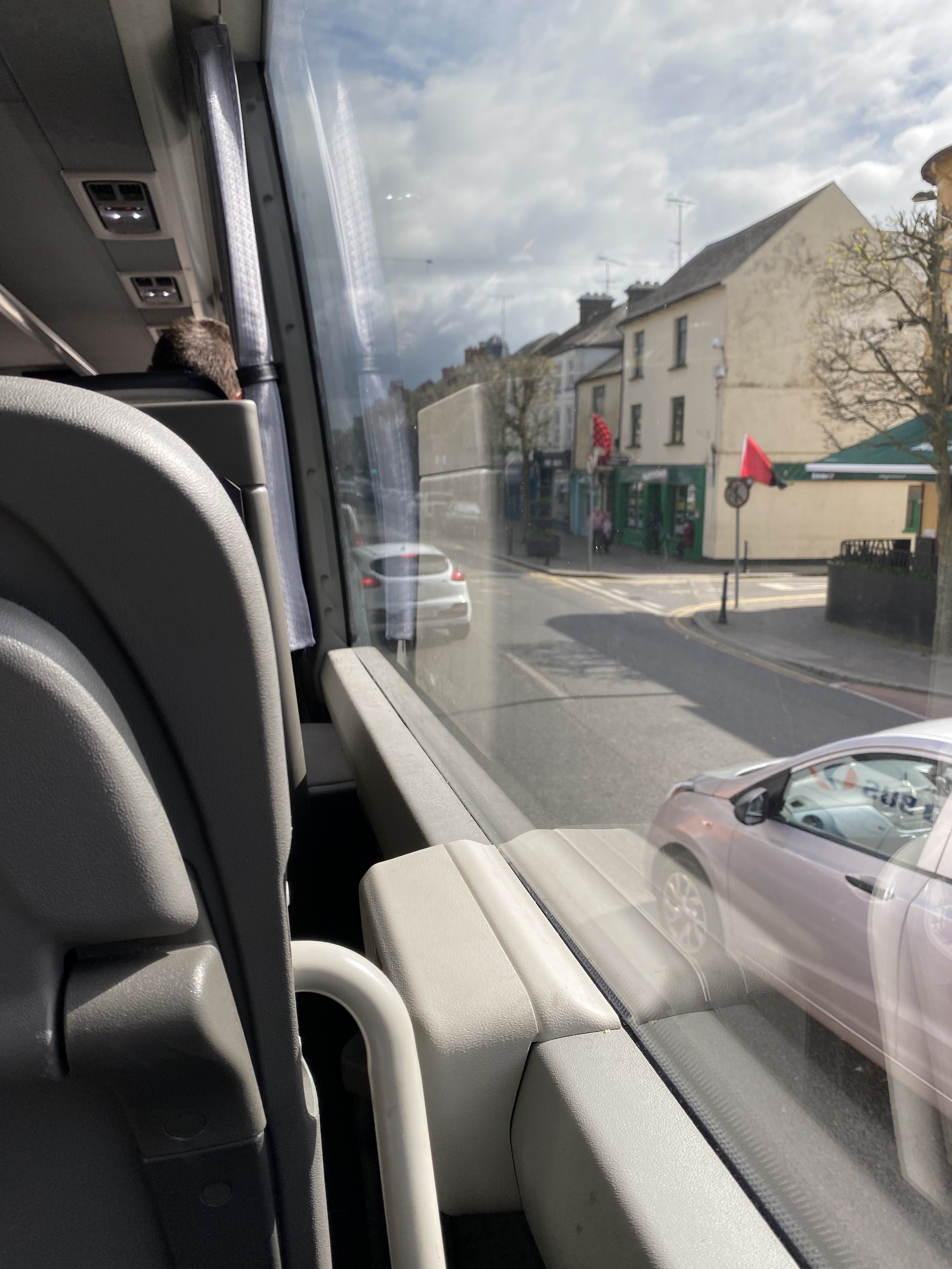 View from bus. Grey interior takes up most of the left while a large window on the right shows a street with cars passing two story shop buildings 