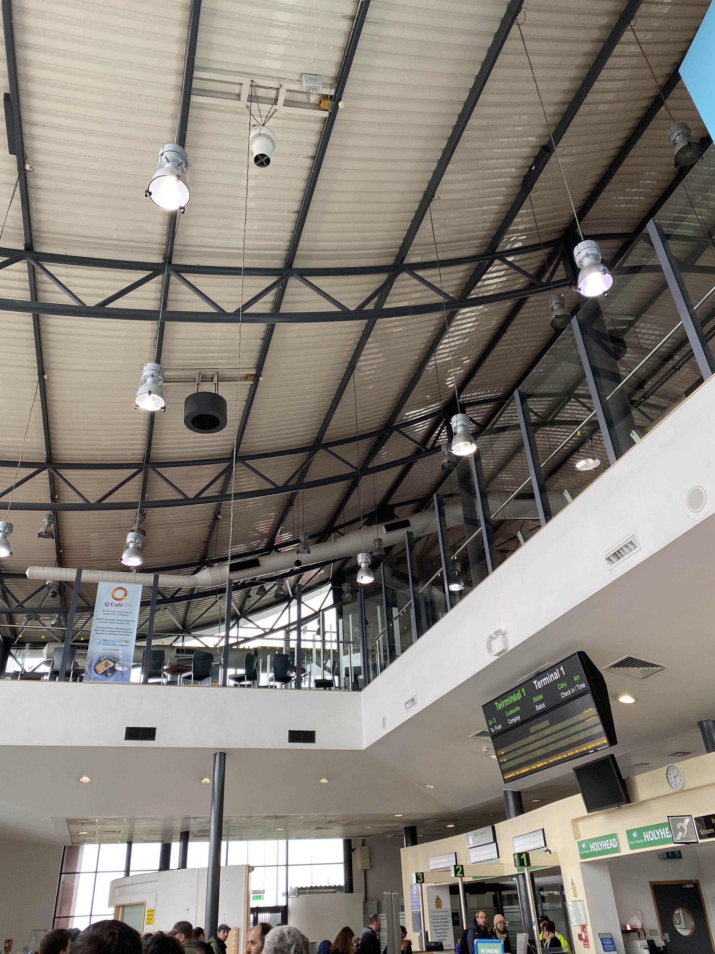 People queueing at the very bottom of the picture. In the middle to the right the check-in desks are visible and a destination sign hangs from the ceiling. The first floor cafe and the sloping terminal roof can be seen to the top.