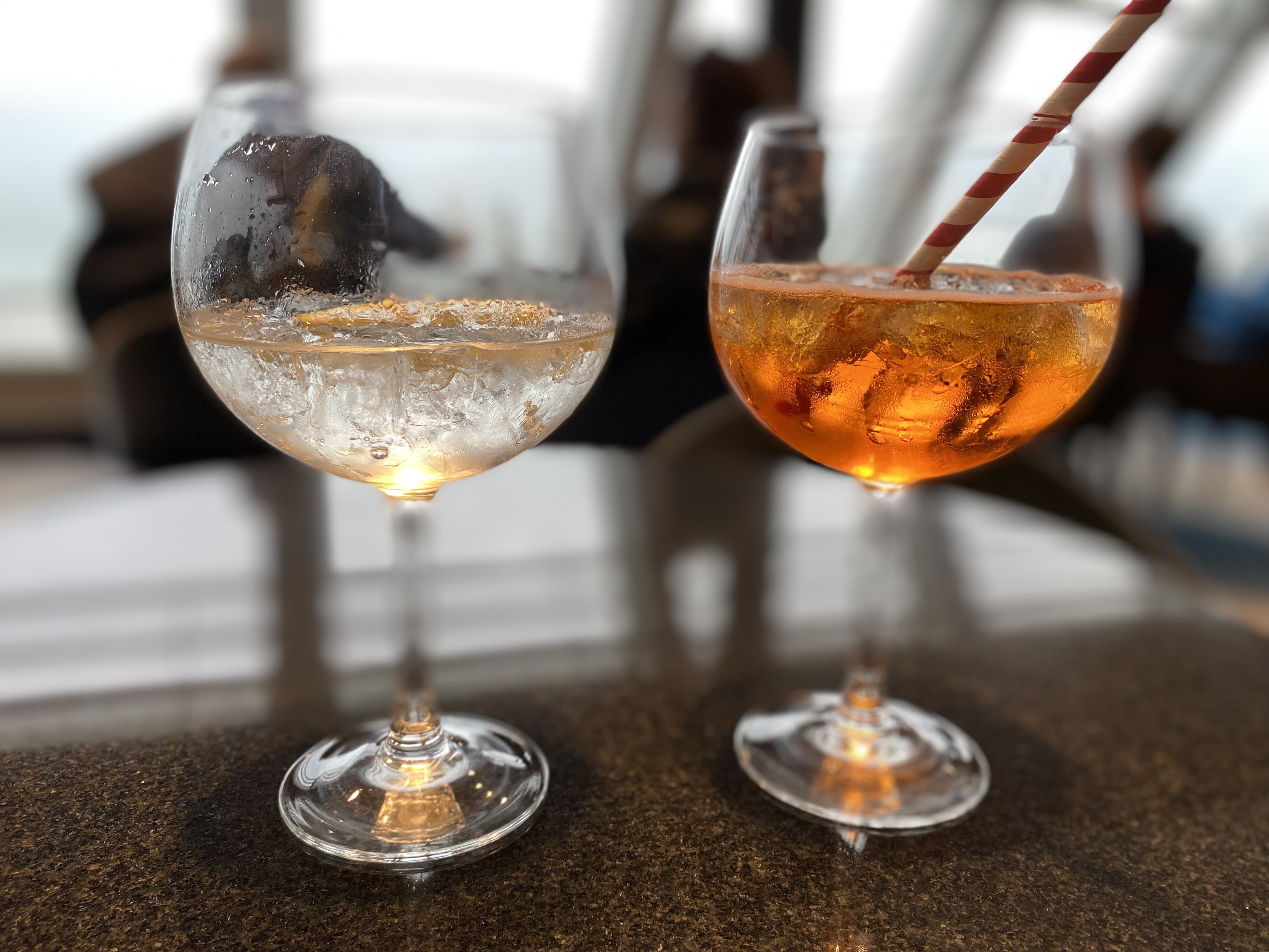 A close-up of a gin and tonic and an apéro spritz with the front windows of a ferry bar out of focus in the background 