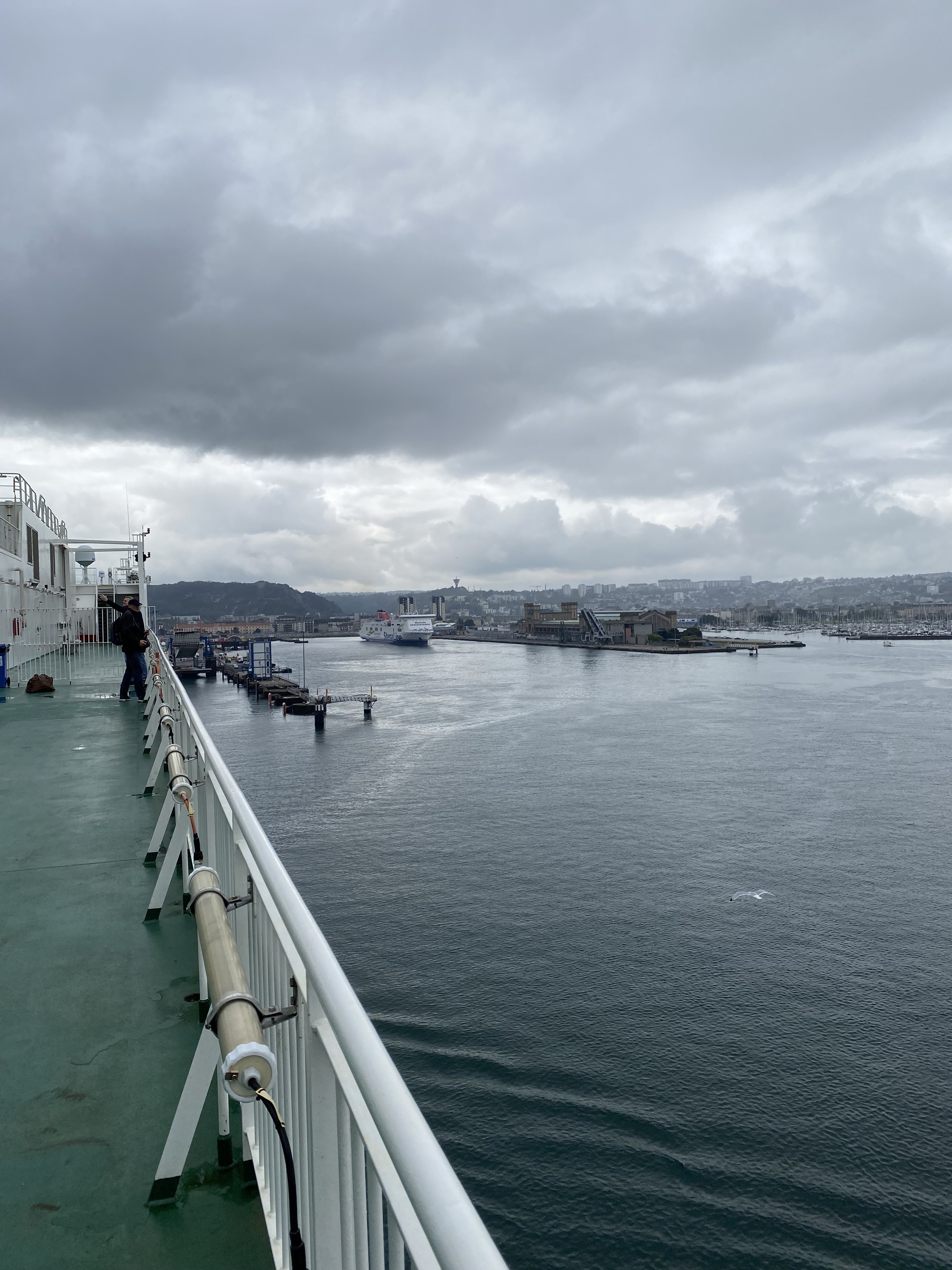 Photo from ferry ship deck. On the left is the green deck and white railing of the ship. On the right the steely grey water of the harbour. A grey sullen sky sits over the port and town. In the distant centre a Stena ferry lies just off its dock