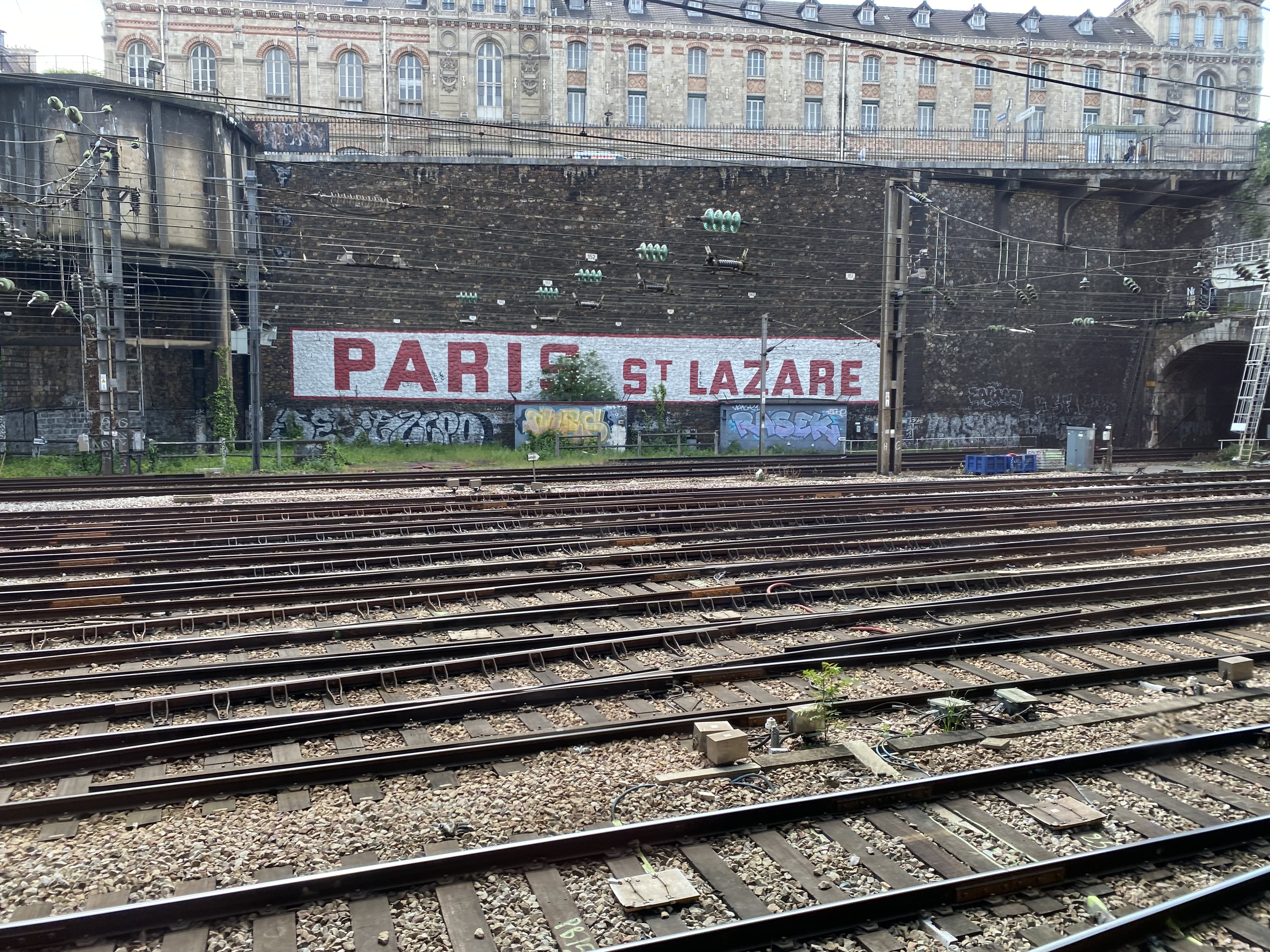 Train tracks and a high wall with mansion buildings above. Paris St Lszare in red lettering on a white background in the centre