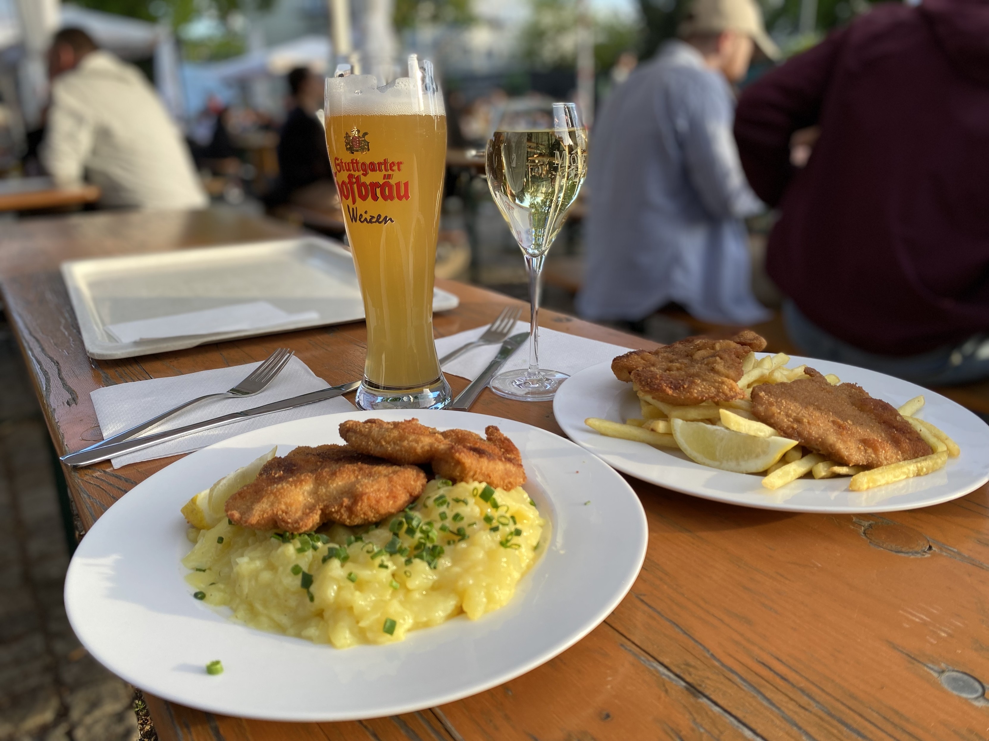 Food and drink at a beer garden. A wheat beer in the traditional high glass and a Riesling in a wine glass, along with two plates of pork schnitzel with potato salad and chips.