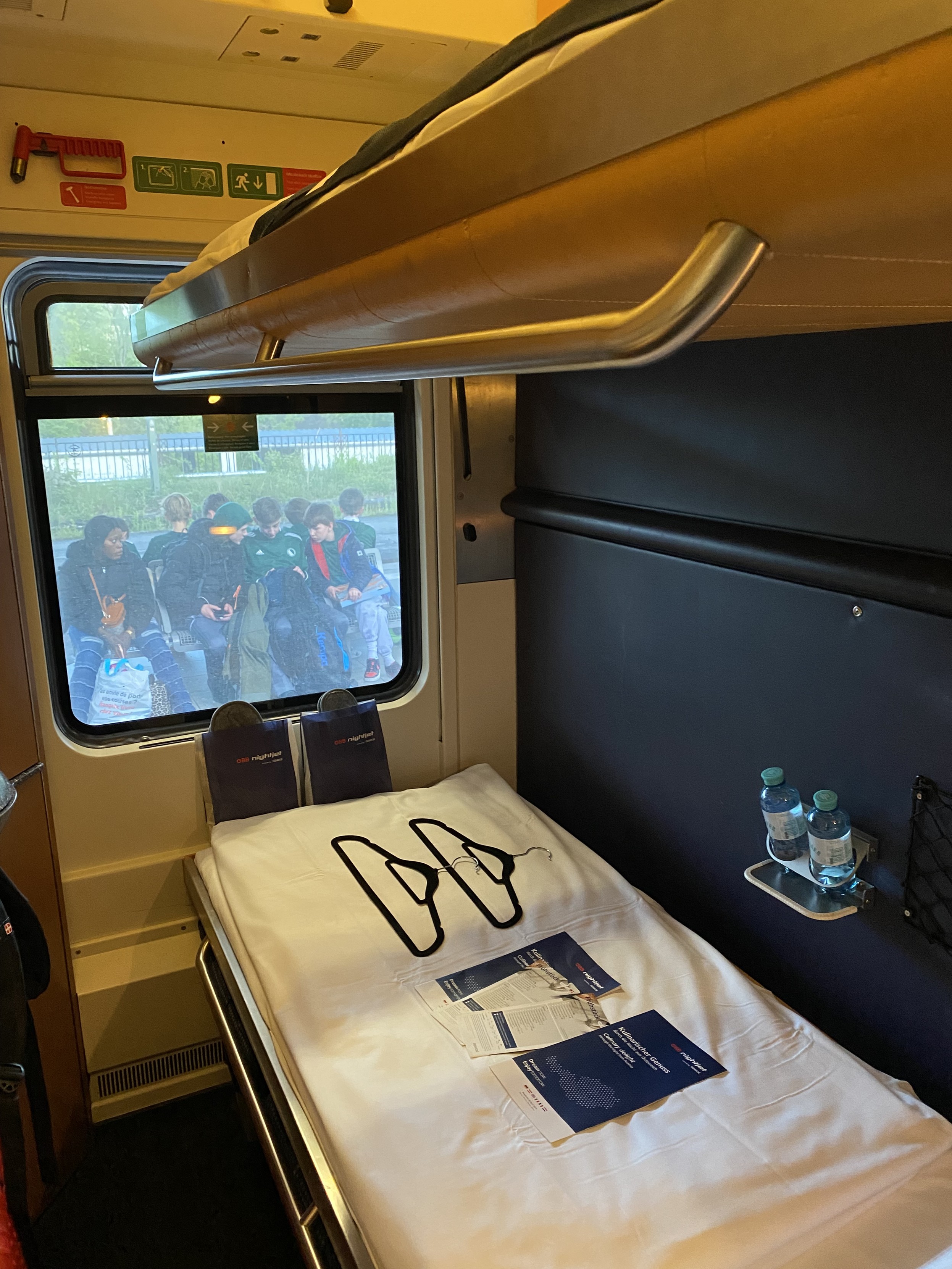 Night train double sleeper cabin. Two bunk beds take up most of the picture. White linen and welcome pack on the lower bunk. The top bunk is just visible. A small window looks out to passengers seated on the platform.