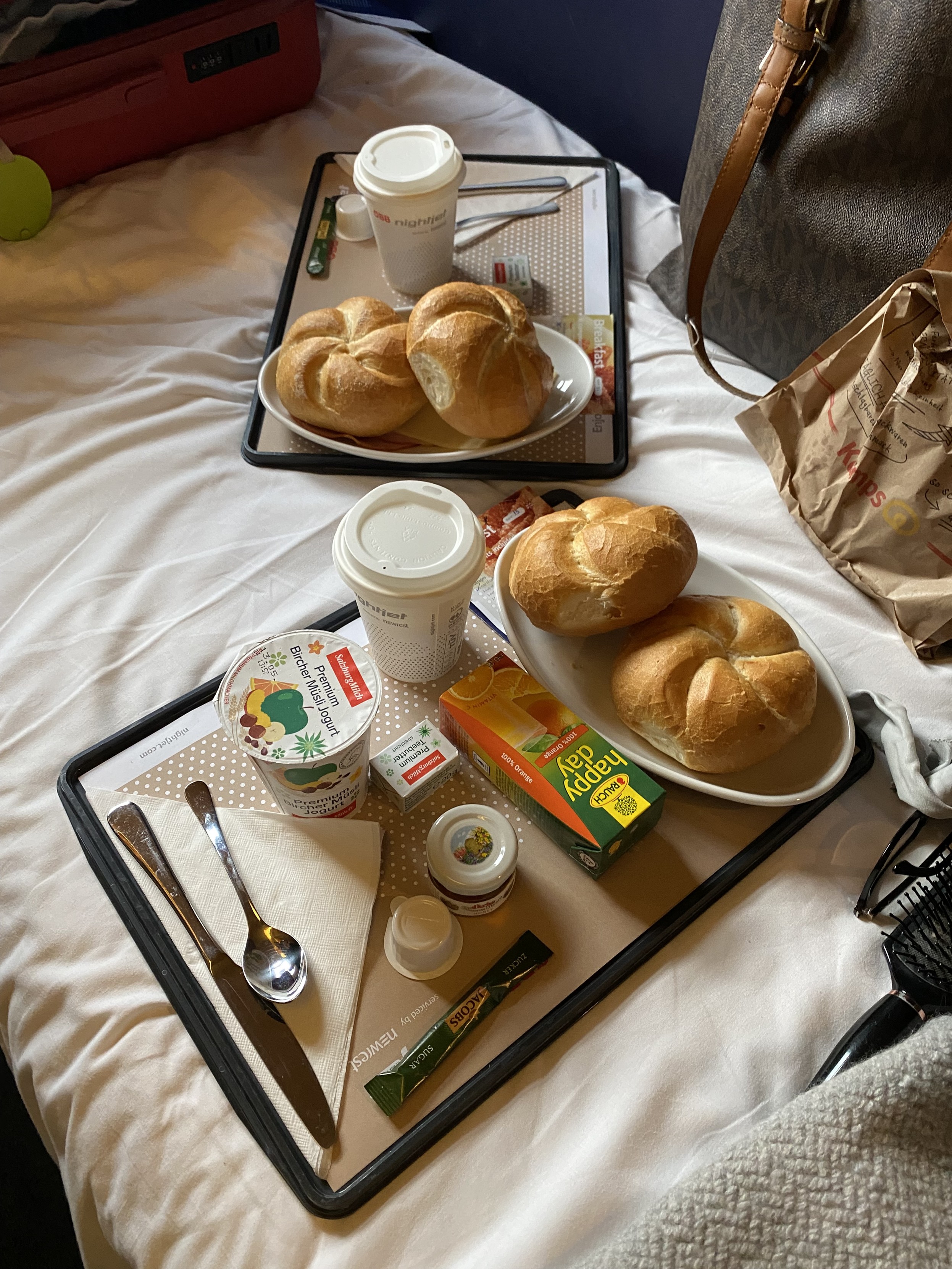 Two breakfast trays on white bed linen surrounded by sundry travel items. Both trays have bread rolls and coffee and cutlery. Orange juice, honey, cheese and ham also present