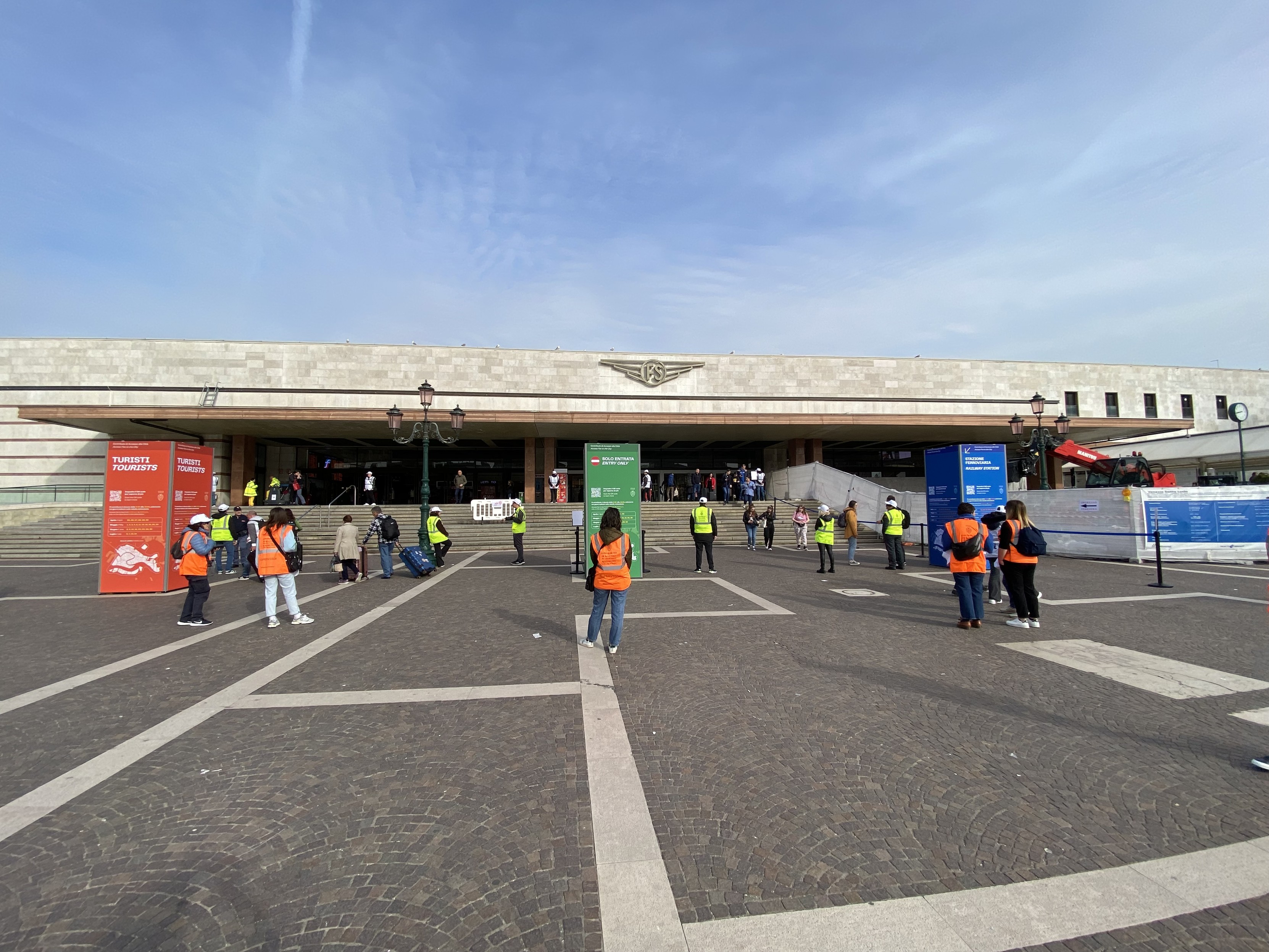 View of the front of Venezia Santa Lucia station. Geometric grey paving in the foreground. Coloured rectangular signs with high viz wearing officials checking passes and the stations monolithic horizontal form in the mid ground. All topped with a milky blue sky