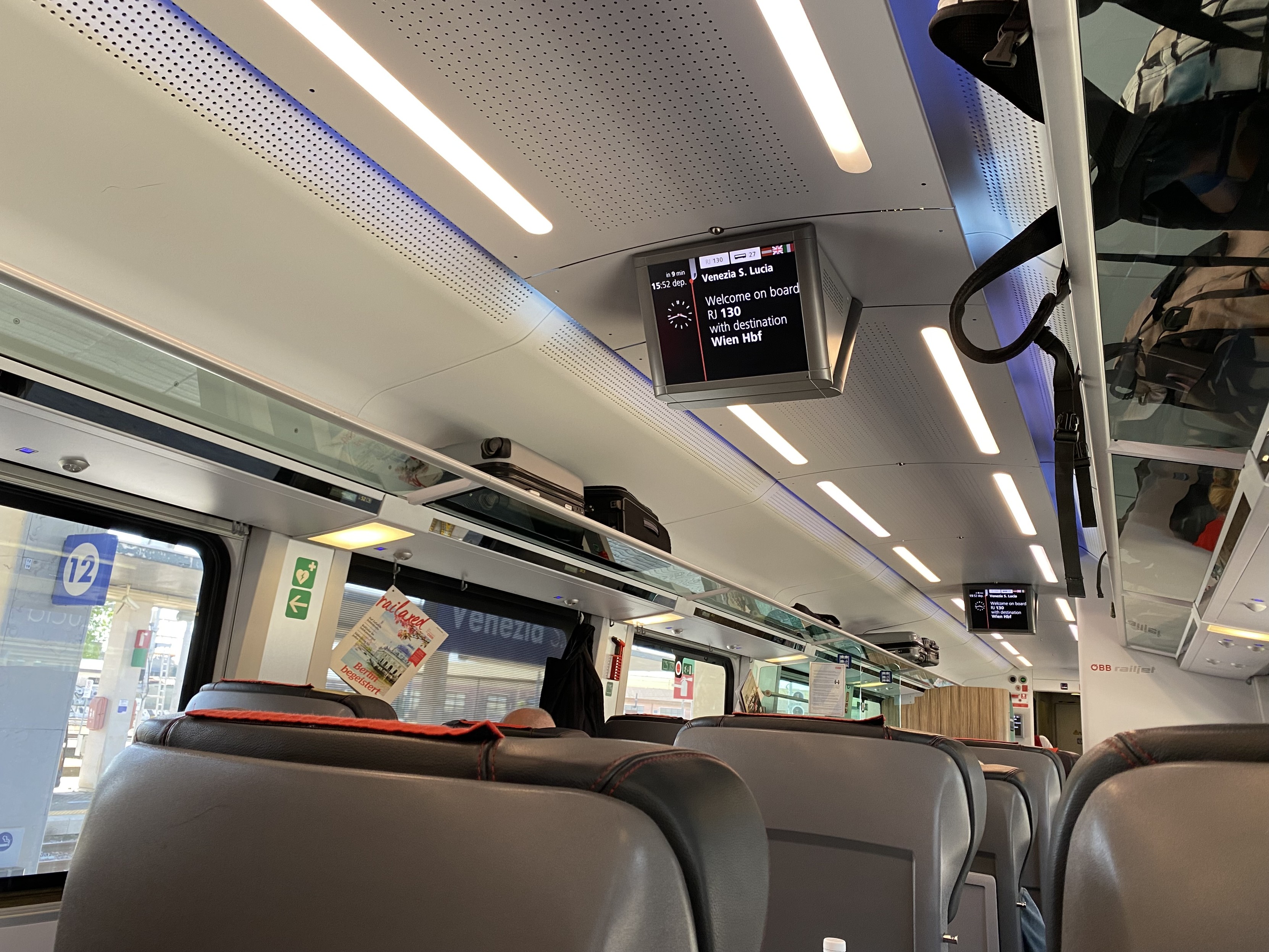 Interior of a Railjet 1st Class carriage. Seats are in a 2+1 arrangement, mostly airline style with just a few facing each other across a table. Large luggage rails are about one third full. The train information sign reads “Venezia S.Lucia, Welcome on board RJ 130 with destination Wien Hbf”