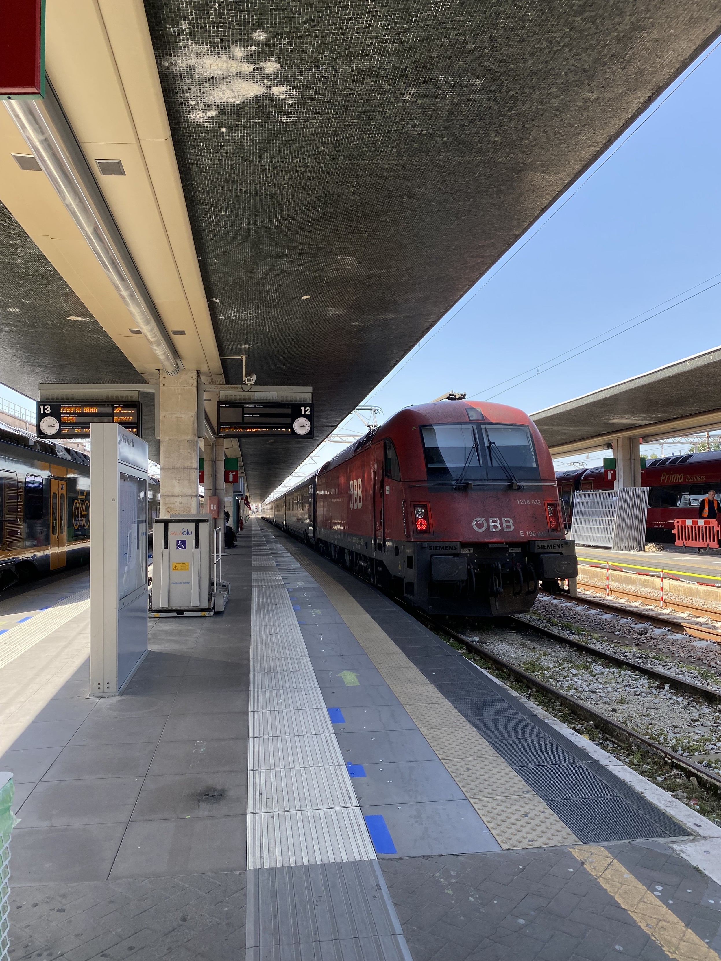 A red train engine at the platform in Venice Santa Lucia station. Large white lettering reads ÖBB. (the letters stand for Österreichische Bundesbahnen or Austrian Federal Railways). The train carriages and platform stretch into the distance.