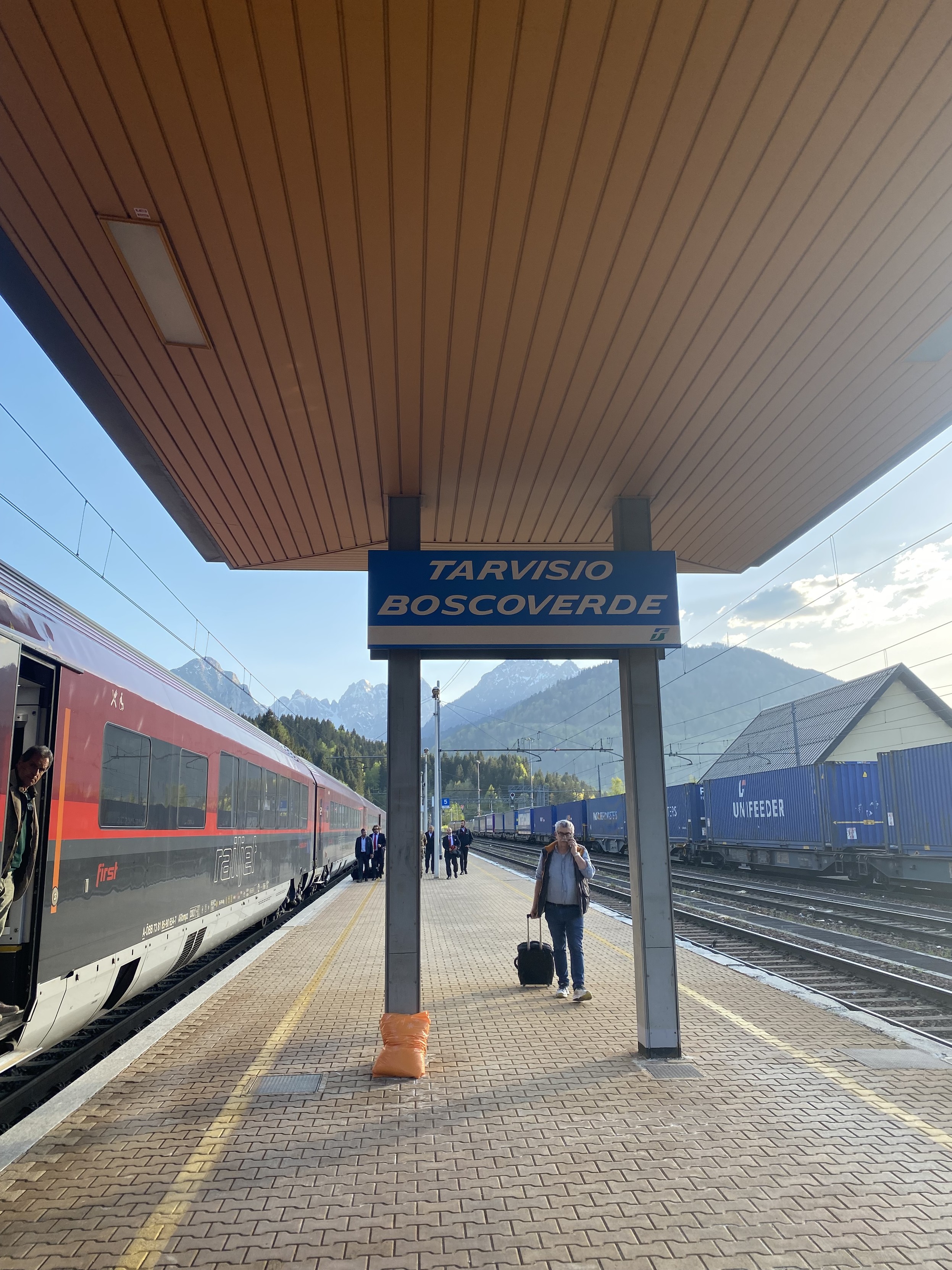 Station platform with a large sign in the centre of picture with white lettering on a blue background. The sign reads Tarvisio Boscoverde. The platform has a white panelled ceiling and its floor is cobble locked grey paving. The red and black train is on the left and glimpses of mountains, buildings, freight carriages, blue ky and white cloud are all to be seen