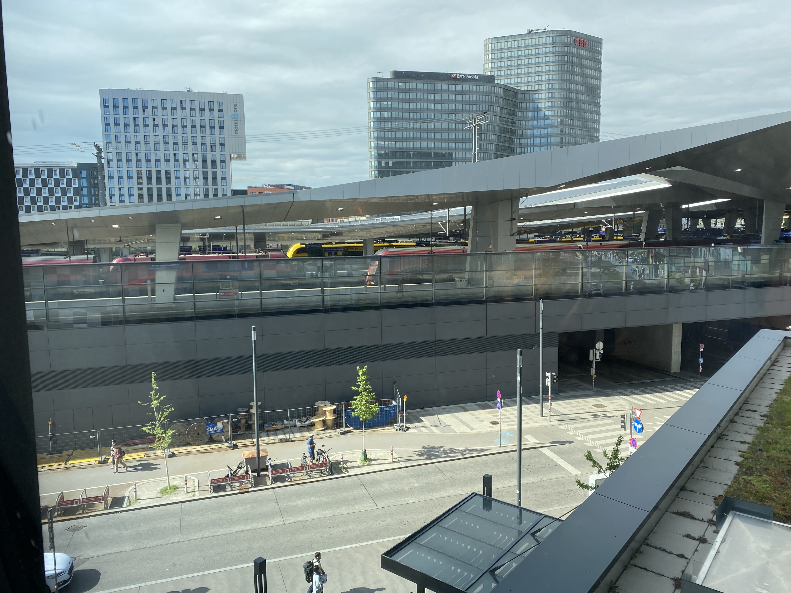 A picture mostly in grey tones. Multiple platforms of the train station are visible with several trains. The angular grey roof rises to right supported by large concrete columns. 10-15  story buildings rise behind backed by a light grey sky. In the foreground a street scene with a few people walking and the entrance to an road underpass of the station