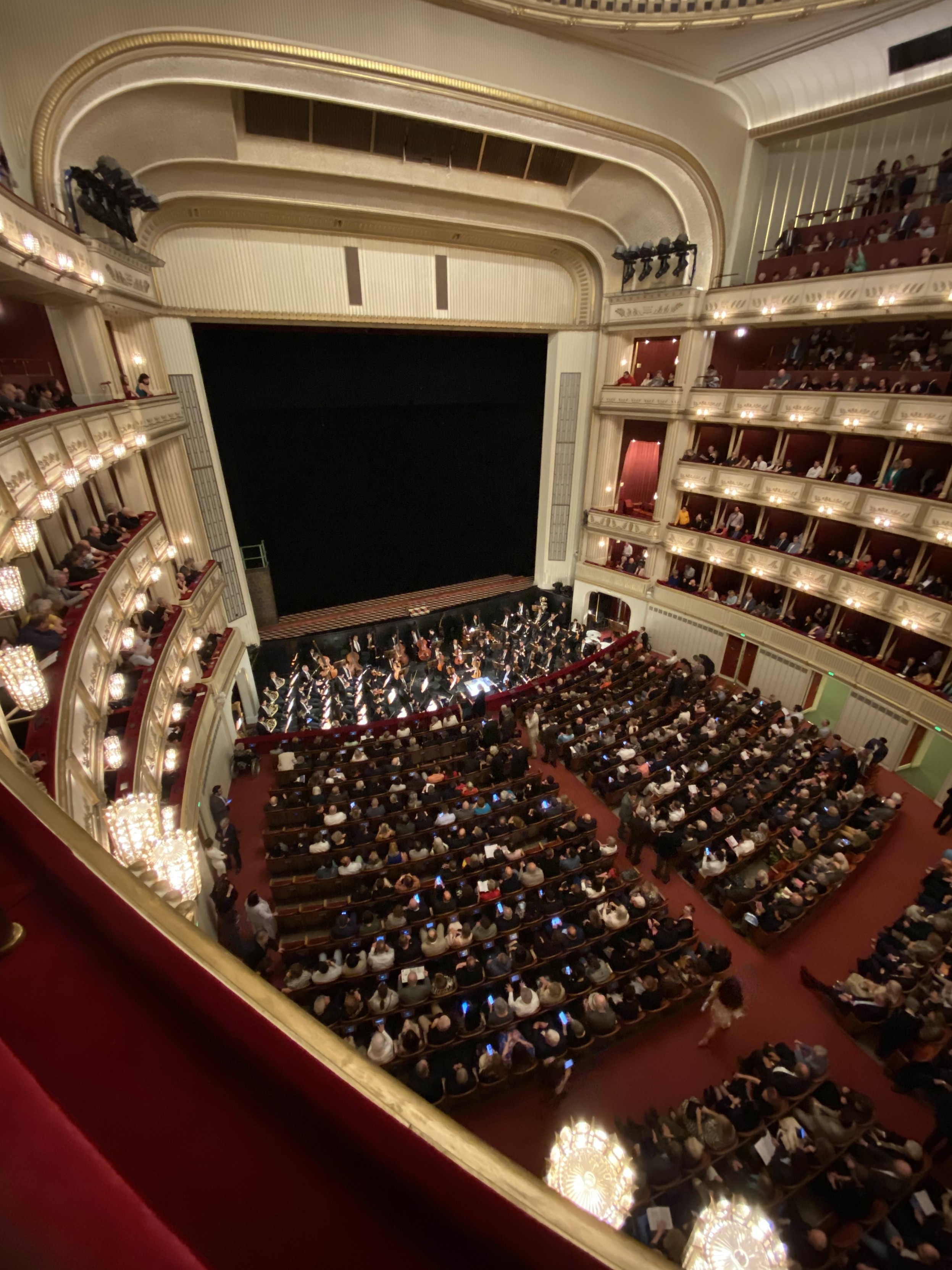 View from an upper balcony in the Wiener Staatsoper. The stage is in darkness in the upper left. The ground floor audience and the orchestra are visible in the centre. Balconies and boxes wrap around on the left and right.