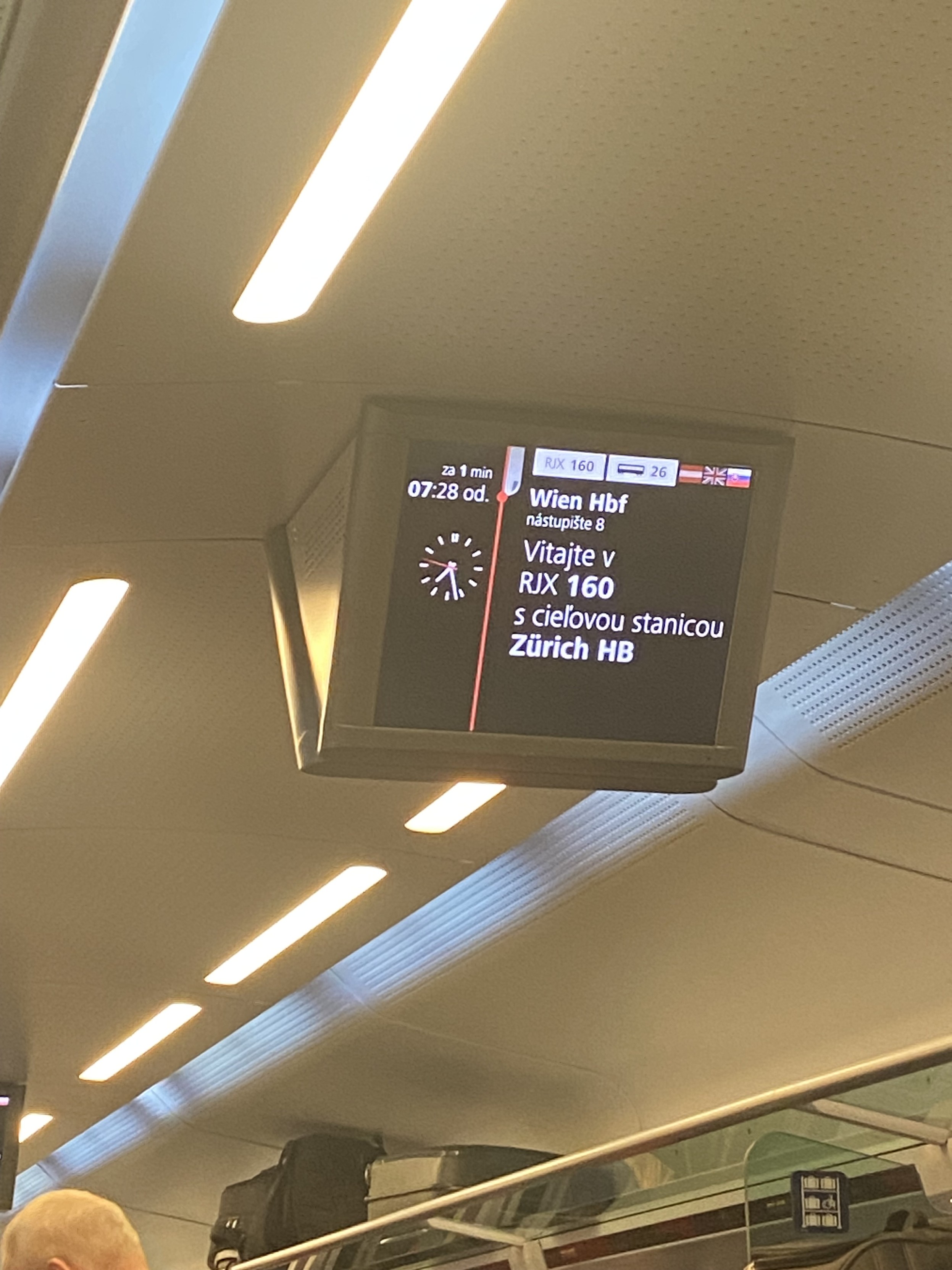 Train information sign. The left third shows departure as “za 1 min 07:28 od.” And an analogue clock showing the current time. The right two thirds displays on the top line: “RJX 160” in dark lettering on a white rectangle, a train coach symbol and “26” on another white rectangle and three flags for the display languages: Austrian, English, Slovak. The following text (in Slovak) is displayed below: “Wien Hbf nastupiste 8 Vitate v RJX 160 s cielovou stanicou Zürich HB”.