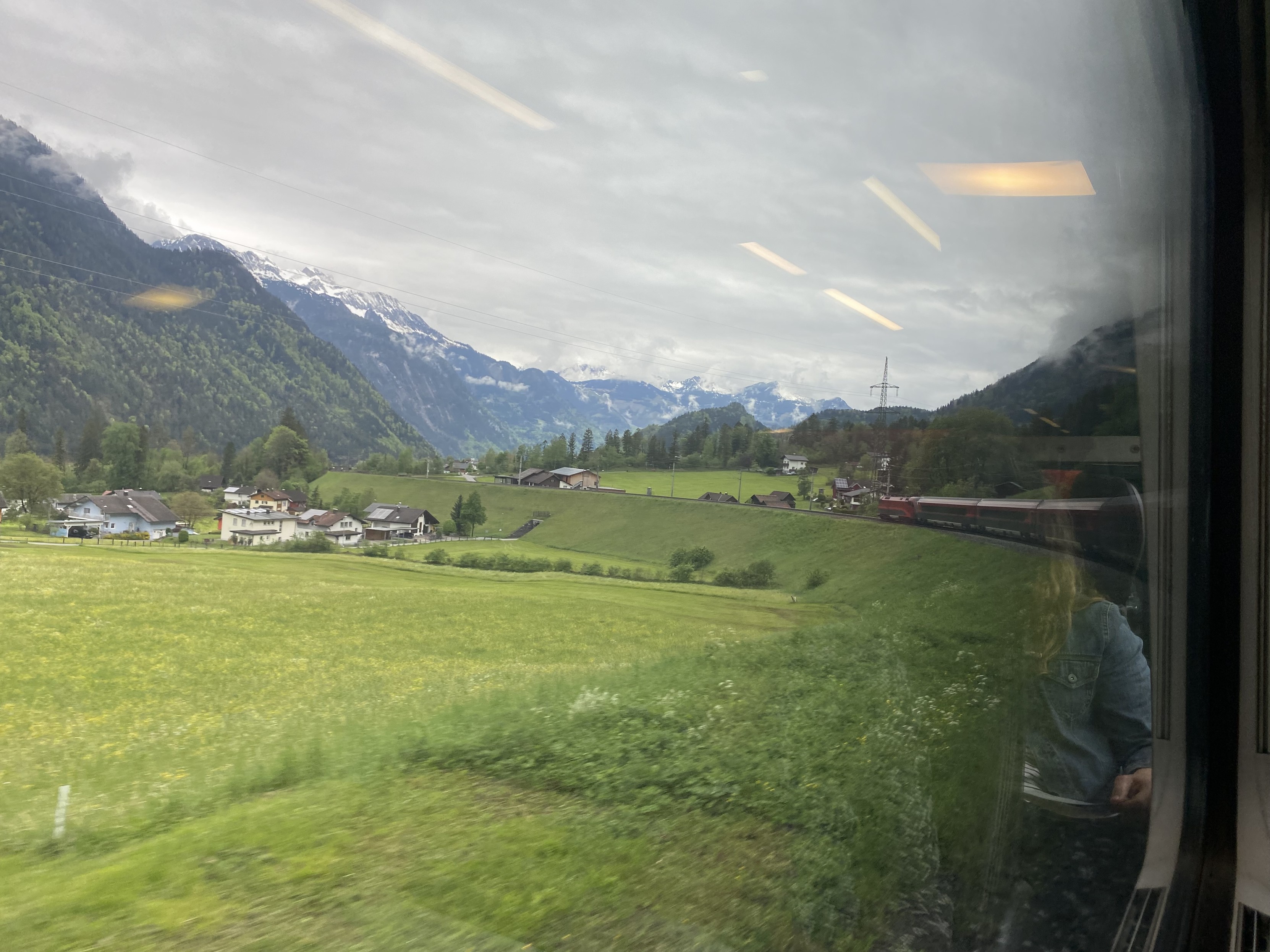 Alpine railway scene. A red ÖBB railjet train follows a curve on the track. Green meadows yield to green hillside to grey and snow capped mountains 