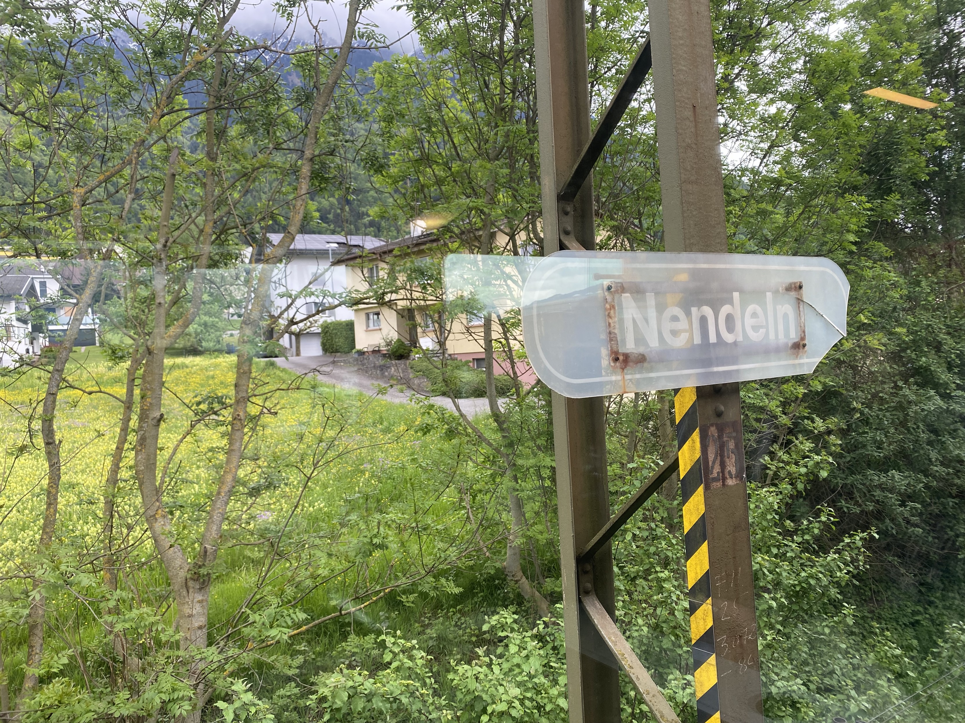 A partially opaque white sign at a countryside railway station reads “Nendeln”. In the background is green vegetation and a cluster of houses