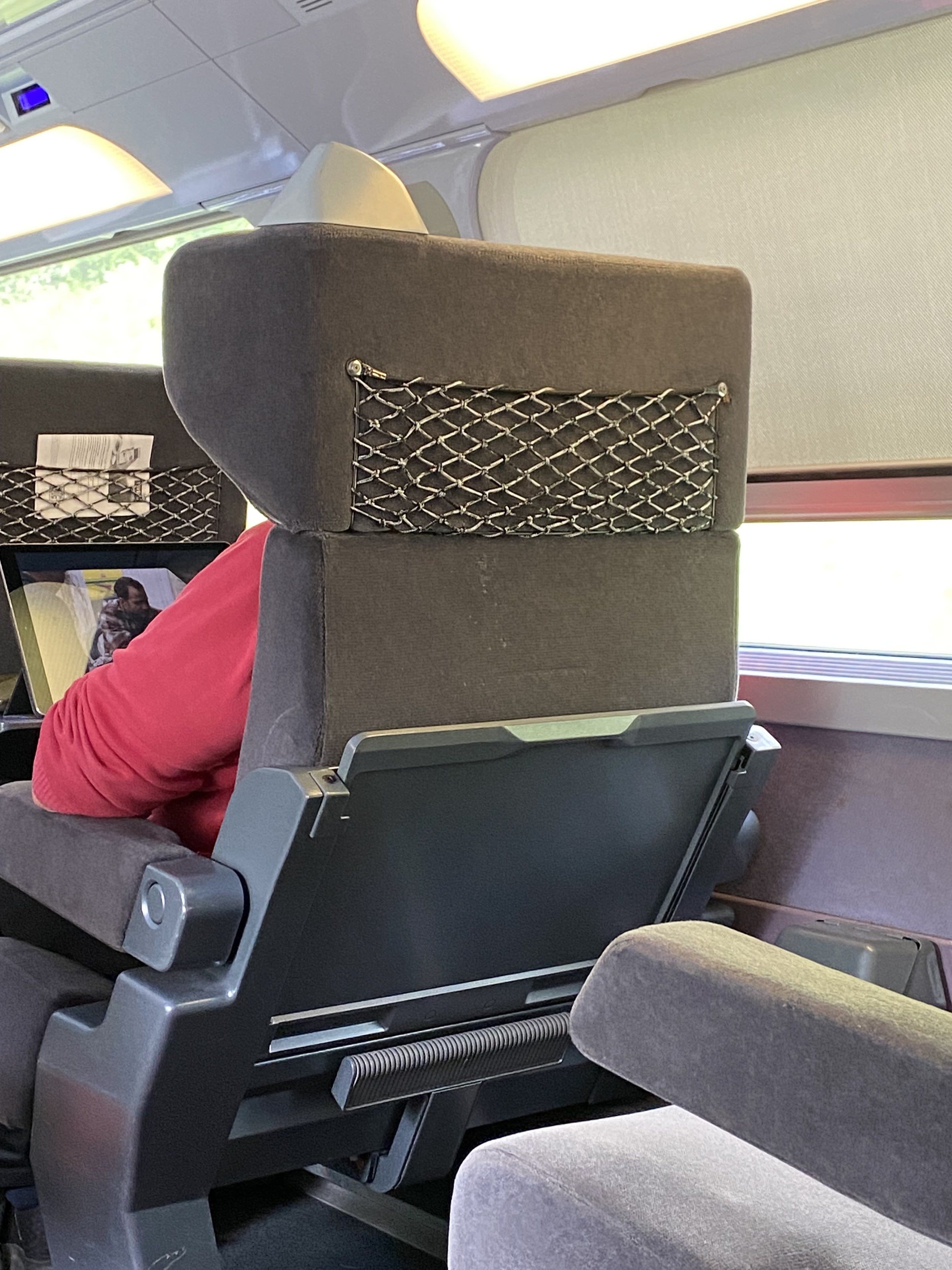 The rear of a grey seat on a train that’s occupied by a person watching a movie on a large screen. The lowered window blind can be seen on their right