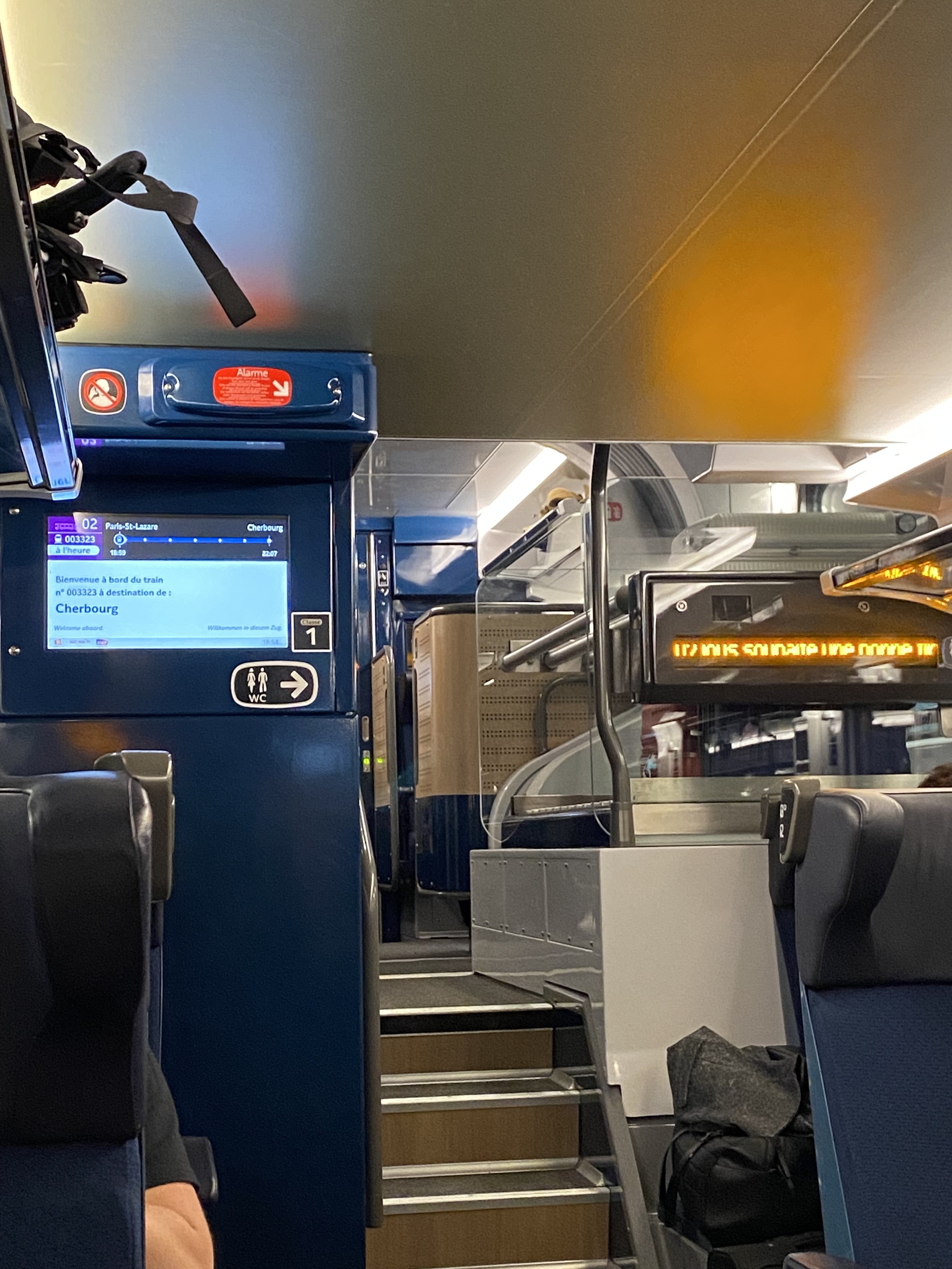 Interior view of a downstairs 1st class carriage of train to Cherbourg. The edges of some seats are visible on the right and left. On the left a screen shows the destination as Cherbourg. On the right a dot matrix display scrolls a message in French. Three steps at the centre lead further down the carriage and on the right a large luggage storage area is visible.