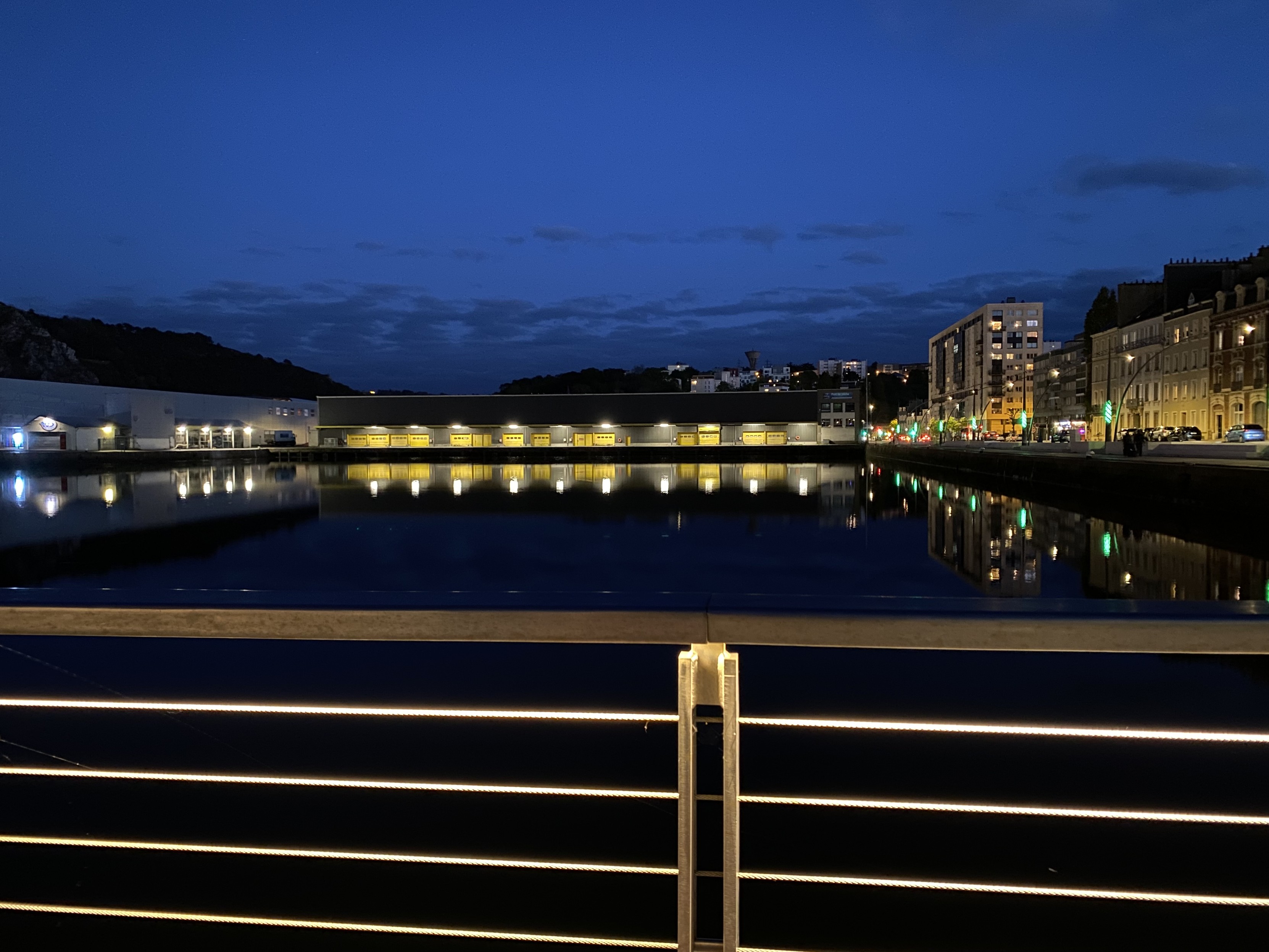 Fishing port in the centre of Cherbourg as night falls. Illuminated horizontal bars of a pedestrian bridge in the foreground. Inky black water in the centre of picture reflecting some harbourside lights. Buildings to both the left and right with the right hand side much brighter. Fishing warehouses mark the end of the harbour - their lights reflected in the water. A mostly clear grey sky with some lower dark grey cloud completes the scene.