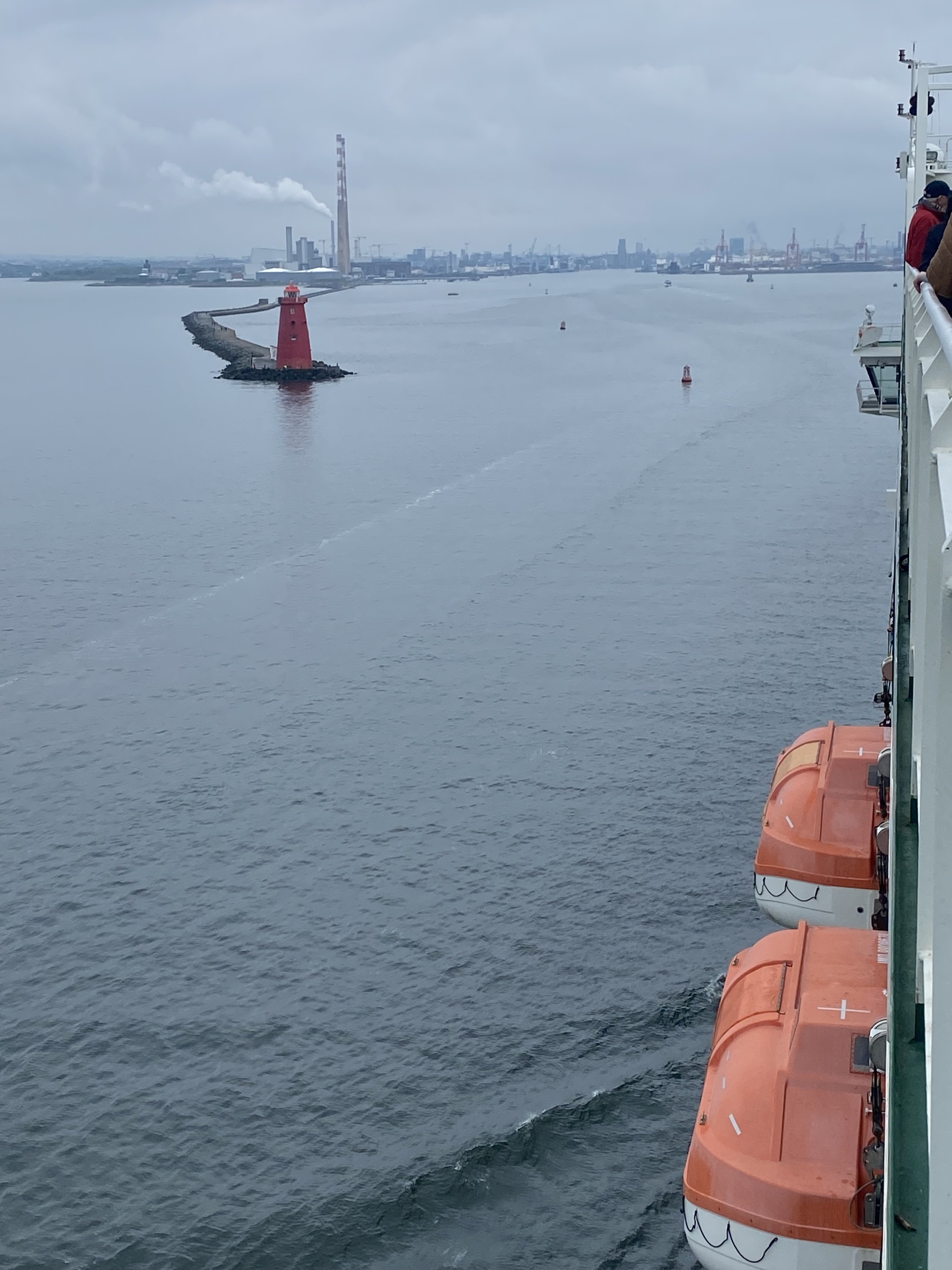 View from ferry deck of the approach into Dublin harbour. Two white and orange life-boats are visible on the side of the ship. Most of the photo is the grey sea with a red lighthouse at the end of the harbour wall. A grey sky hangs over the grey horizontal line of the cityscape punctured by the Poolbeg chimneys