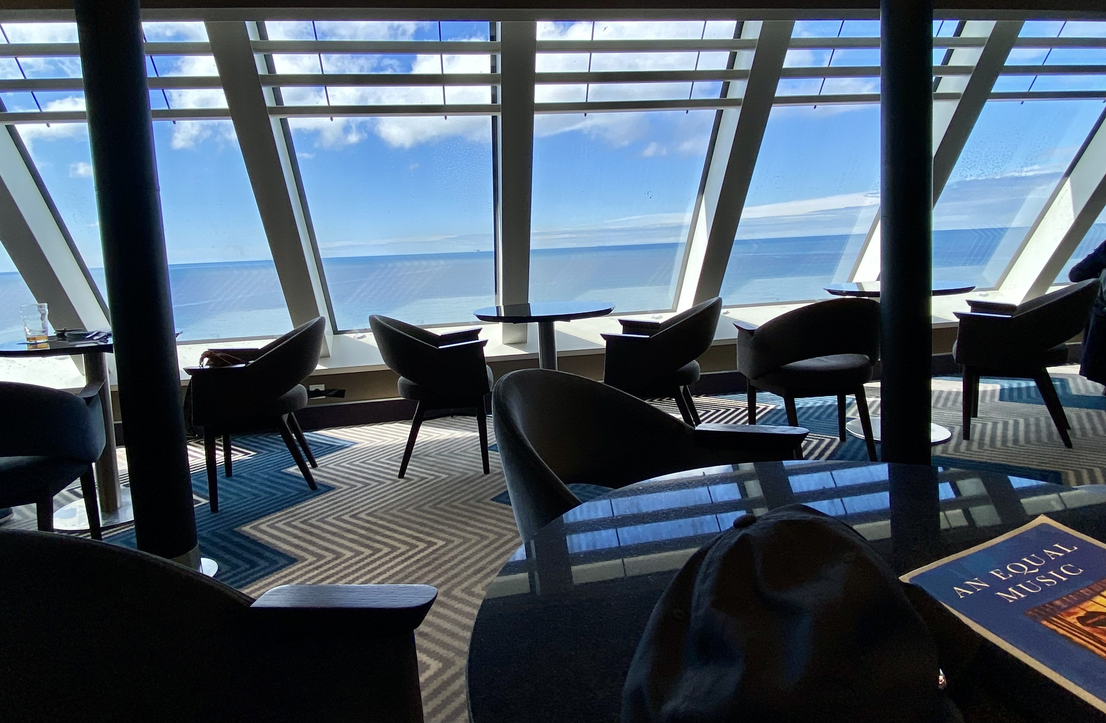 A sunny view from the bar at front of the Irish Ferries WB Yeats. Bar tables and chairs in the foreground and a blue sea and sky are visible through large white framed windows 